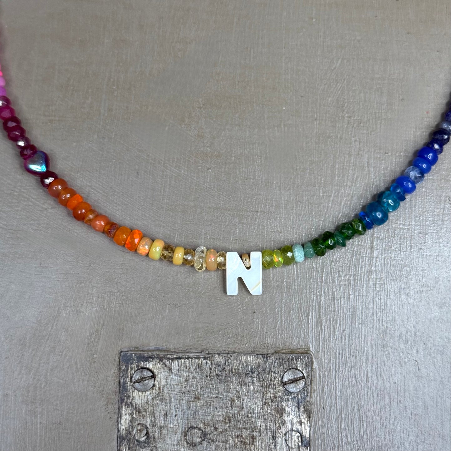 Necklace with rainbow gemstone beads and a mother of pearl 'N' pendant on a textured surface.