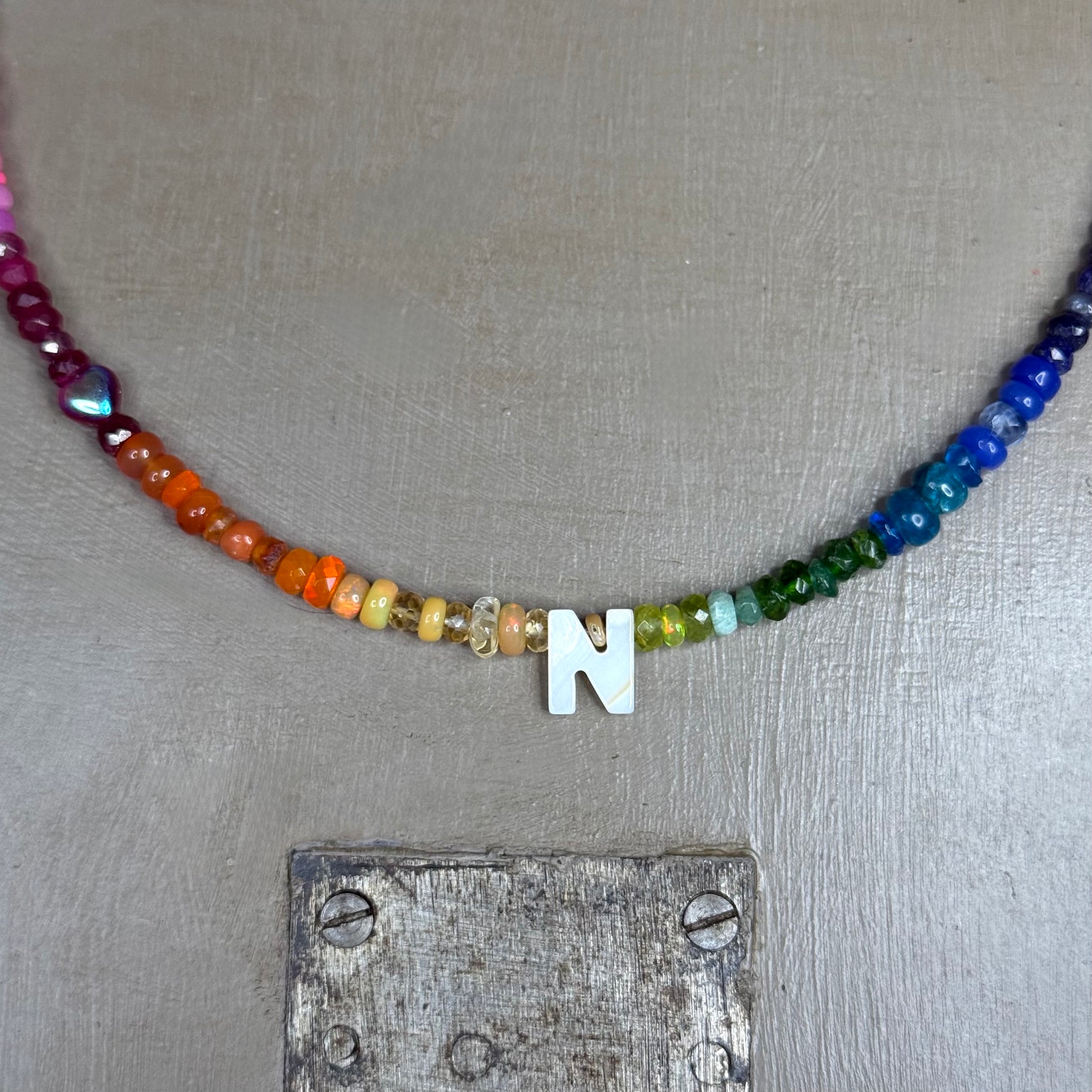 Necklace with rainbow gemstone beads and a mother of pearl 'N' pendant on a textured surface.