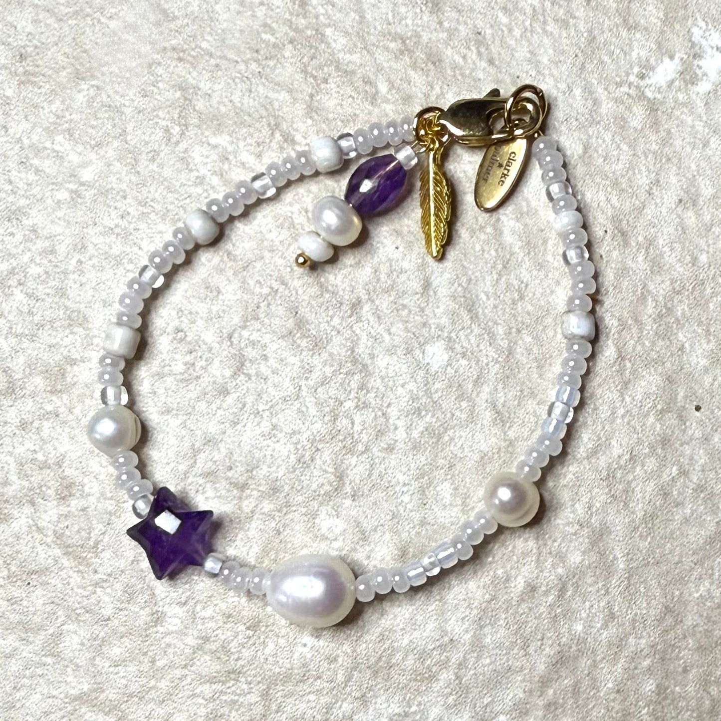 Pearl bracelet with purple amethyst star charm and gold feather charm on a travertine  surface