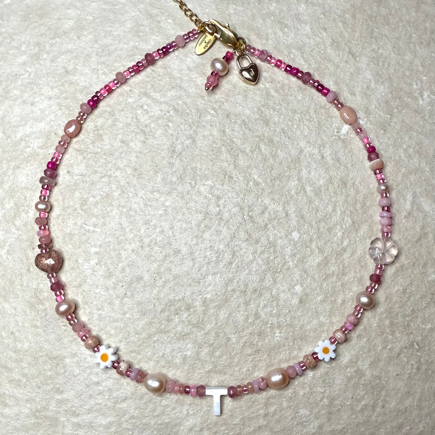 Detail of Necklace with pink gemstone beads and a letter 'T' on a textured white background